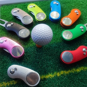 Könnte beinhalten: Sortiment an bunten Golfballmarkern mit einem weißen Golfball auf grünem Untergrund. Die Marker sind aus Metall und haben verschiedene Designs.