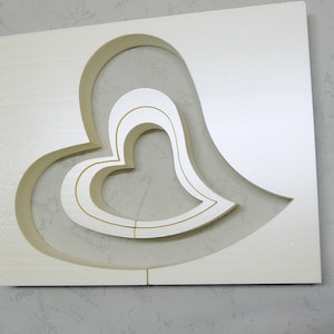 Könnte beinhalten: Eine weiße Schaumstoffplatte mit drei konzentrischen herzförmigen Ausschnitten. Die Ausschnitte sind übereinander gelegt und erzeugen einen dreidimensionalen Effekt.