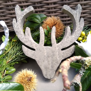 Può includere: Un ornamento per la testa di renna in cemento grigio circondato da vegetazione, tra cui rami di pino, piccoli fiori bianchi e baccelli di semi marroni e spinosi.