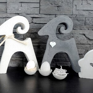 Può includere: Decorazioni pasquali in cemento. Due sculture di arieti, un coniglietto e tre piccole ciotole con uova e nidi. Gli arieti sono bianchi e grigi, con un ciondolo a forma di cuore su uno. Il coniglietto è bianco.