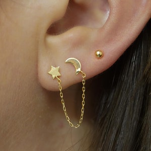 May include: Gold star and crescent moon earrings connected by a delicate gold chain. The earrings are worn in the upper lobe of the ear.