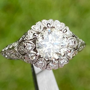 May include: A silver engagement ring with a large, round, clear diamond surrounded by a halo of smaller diamonds. The band is decorated with intricate floral designs.