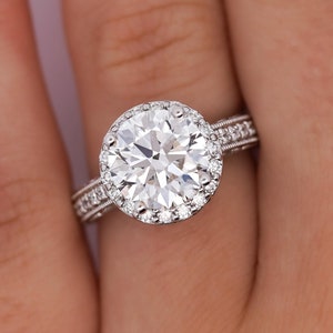 May include: A silver engagement ring featuring a large round diamond centre stone encircled by a halo of smaller diamonds. The ring has a delicate band with a milgrain edge.