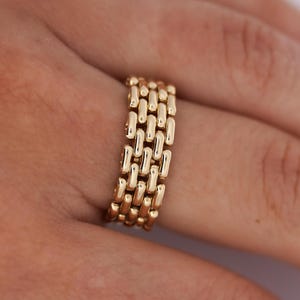 Op de afbeelding: Een goudkleurige ring met een schakelkettingontwerp. De ring is breed en heeft een reeks rechthoekige schakels die in rijen zijn gerangschikt. De ring wordt aan een vinger gedragen, met de hand op de achtergrond.