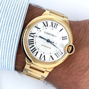May include: A gold Cartier watch with a round white face and Roman numeral hour markers. The watch has a date window at the 12 o'clock position and the words 'Cartier' and 'Automatic' printed on the face.