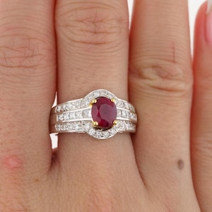 Può includere: Un anello d'oro con un grande rubino ovale circondato da un alone di diamanti più piccoli. L'anello ha un design a tripla fascia con diamanti incastonati nelle fasce.