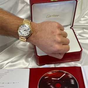 May include: A gold Cartier watch with a white face and Roman numeral hour markers. The watch is in a red box with the Cartier logo on the top.