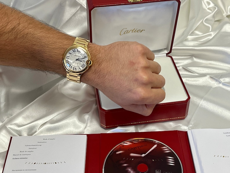 May include: A gold Cartier watch with a white face and Roman numeral hour markers. The watch is in a red box with the Cartier logo on the top.