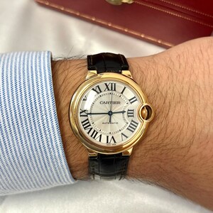 May include: A gold Cartier watch with a black leather band. The watch face is white with black Roman numeral hour markers and black hands. The watch face also has the words 'Cartier' and 'Automatic' printed on it.