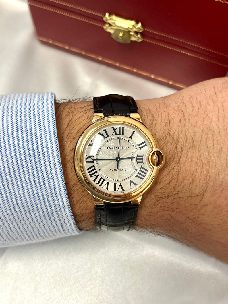 May include: A gold Cartier watch with a black leather band. The watch face is white with black Roman numeral hour markers and black hands. The watch face also has the words 'Cartier' and 'Automatic' printed on it.
