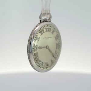 May include: A silver pocket watch with a white face and diamond-encrusted Roman numeral hour markers. The watch face also features the text "Vacheron & Constantin Geneve".