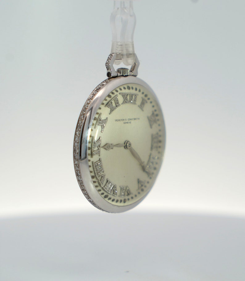 May include: A silver pocket watch with a white face and diamond-encrusted Roman numeral hour markers. The watch face also features the text "Vacheron & Constantin Geneve".