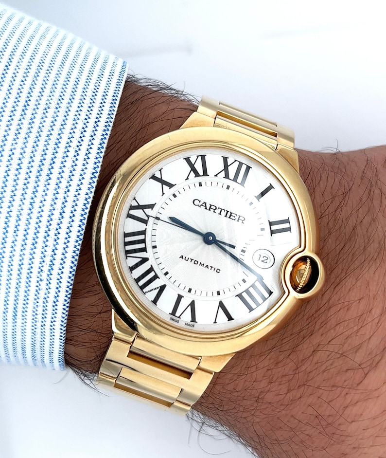 May include: A gold Cartier watch with a round white face and Roman numeral hour markers. The watch has a date window at the 12 o'clock position and the words 'Cartier' and 'Automatic' printed on the face.