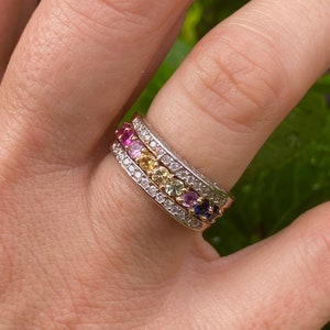 May include: A gold band ring with a row of colorful gemstones set in a white metal band. The gemstones are arranged in a rainbow pattern, with pink, red, orange, yellow, green, blue, and purple stones.