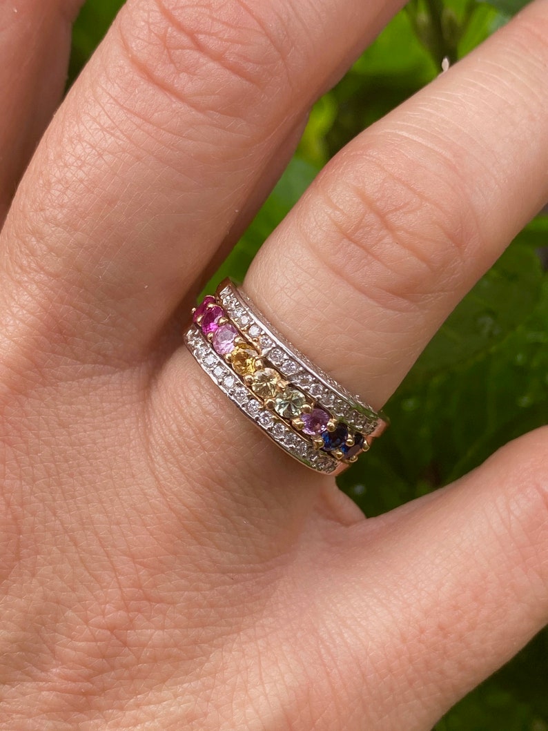 May include: A gold band ring with a row of colorful gemstones set in a white metal band. The gemstones are arranged in a rainbow pattern, with pink, red, orange, yellow, green, blue, and purple stones.