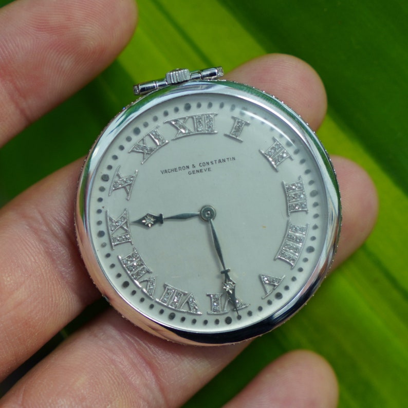 May include: A silver pocket watch with a white face and Roman numeral hour markers. The watch is set with diamonds around the edge of the face and around the hour markers. The watch face also includes the text "VACHERON & CONSTANTIN GENEVE".