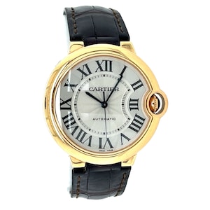 May include: A gold Cartier Ballon Bleu de Cartier watch with a white dial and black Roman numeral hour markers. The watch has a black leather strap and a date window at the 3 o'clock position. The watch face also says 'Automatic' and 'Swiss Made'.