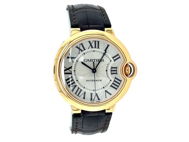 May include: A gold Cartier Ballon Bleu de Cartier watch with a white dial and black Roman numeral hour markers. The watch has a black leather strap and a date window at the 3 o'clock position. The watch face also says 'Automatic' and 'Swiss Made'.