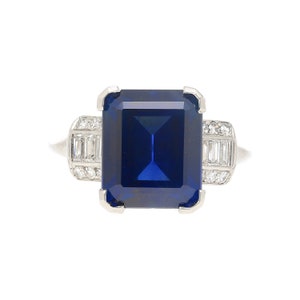 K&ouml;nnte beinhalten: Ein Platinring mit einem gro&szlig;en, tiefblauen Smaragdschliff-Saphir und kleineren Diamanten, die in die Ringschiene eingefasst sind.