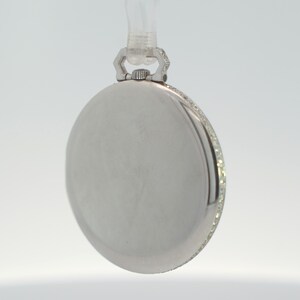 May include: A silver pocket watch with a diamond-encrusted bezel. The watch has a round face and a hinged lid.