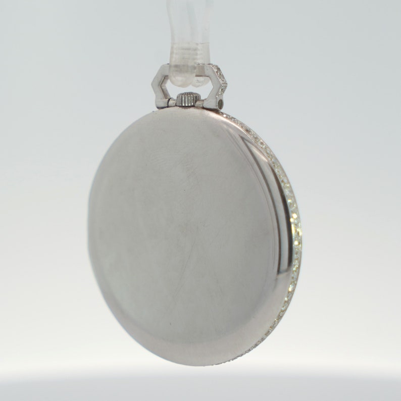 May include: A silver pocket watch with a diamond-encrusted bezel. The watch has a round face and a hinged lid.