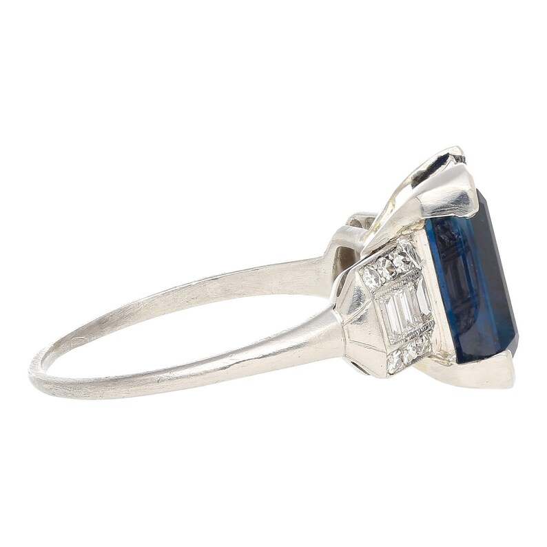 K&ouml;nnte beinhalten: Ein Platinring mit einem gro&szlig;en, tiefblauen Saphir, der von kleineren Diamanten umgeben ist. Der Ring hat eine schlichte Band und ein klassisches Design.