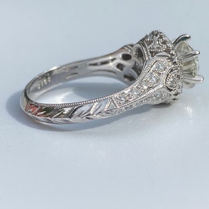 May include: A silver engagement ring with a large center diamond and smaller diamonds set in a halo around it. The band is engraved with a floral design.