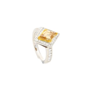 May include: A silver ring featuring a square-shaped yellow gemstone set within a halo of smaller diamonds. The band of the ring is also adorned with diamonds.