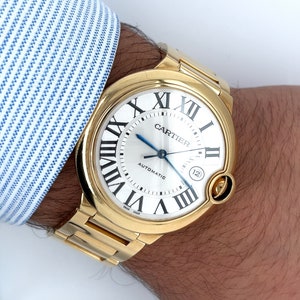 May include: A gold Cartier watch with a round face and Roman numeral markings. The watch face is white with black markings and the words 'Cartier' and 'Automatic' printed on it. The watch has a gold band.