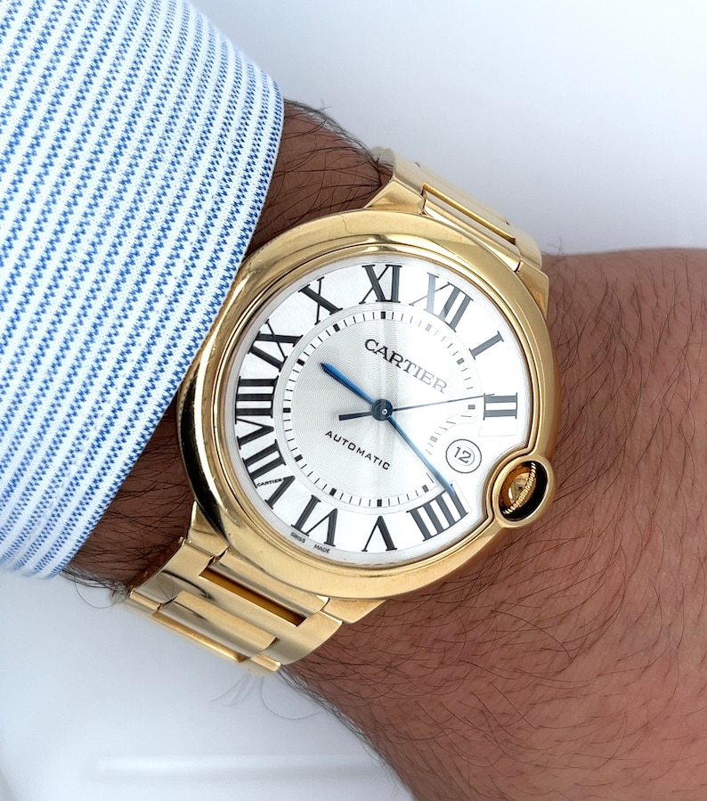 May include: A gold Cartier watch with a round face and Roman numeral markings. The watch face is white with black markings and the words 'Cartier' and 'Automatic' printed on it. The watch has a gold band.