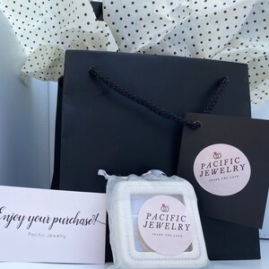 May include: A black gift bag with a white tissue paper lining. The bag has a white tag with the text "Pacific Jewelry Share the Love" and a white box with the same text. The box is sitting on a white surface. The text "Enjoy your purchase! Pacific Jewelry" is on a white card.