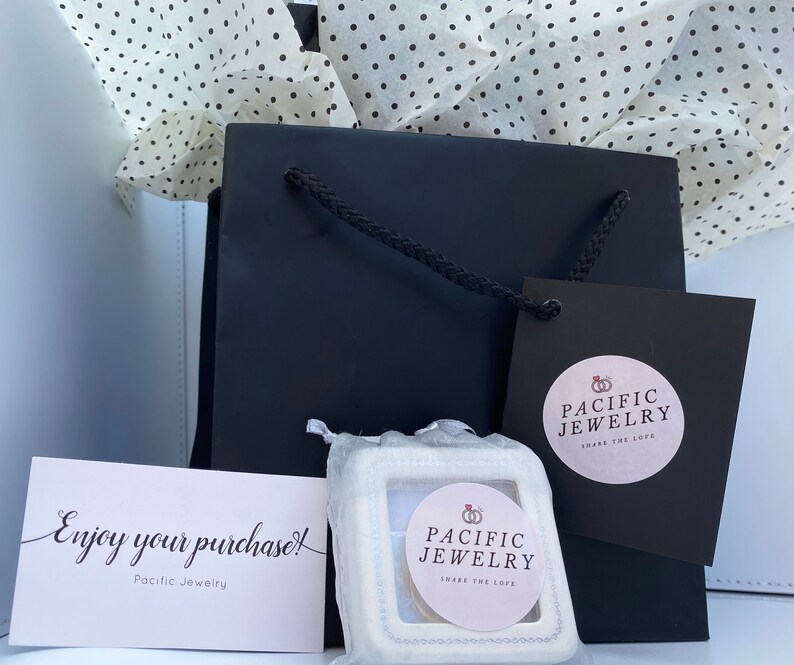 May include: A black gift bag with a white tissue paper lining. The bag has a white tag with the text "Pacific Jewelry Share the Love" and a white box with the same text. The box is sitting on a white surface. The text "Enjoy your purchase! Pacific Jewelry" is on a white card.
