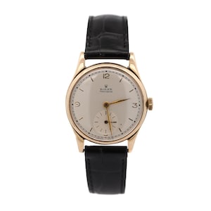 Op de afbeelding: Een goudkleurig Rolex Precision polshorloge met een crèmekleurige wijzerplaat en een zwarte leren band. Het horloge heeft gouden wijzers en uurmarkeringen, met het nummer 12 bovenaan en een kleine secondenwijzer. Een klassiek ontwerp.