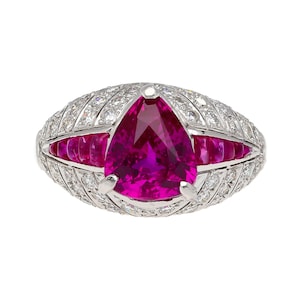 Puede incluir: Un anillo de rubí en forma de pera engastado en oro blanco con un halo de diamantes y una fila de rubíes a cada lado.