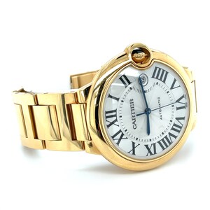 May include: A gold Cartier watch with a round face and Roman numeral hour markers. The watch has a white dial with a blue second hand and the word 'Automatic' printed on it. The watch is on a gold metal bracelet.