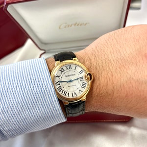 May include: A gold Cartier watch with a white face and black leather band. The watch face has Roman numeral hour markers and a date window at the 3 o'clock position. The watch is labeled 'CARTIER AUTOMATIC'.