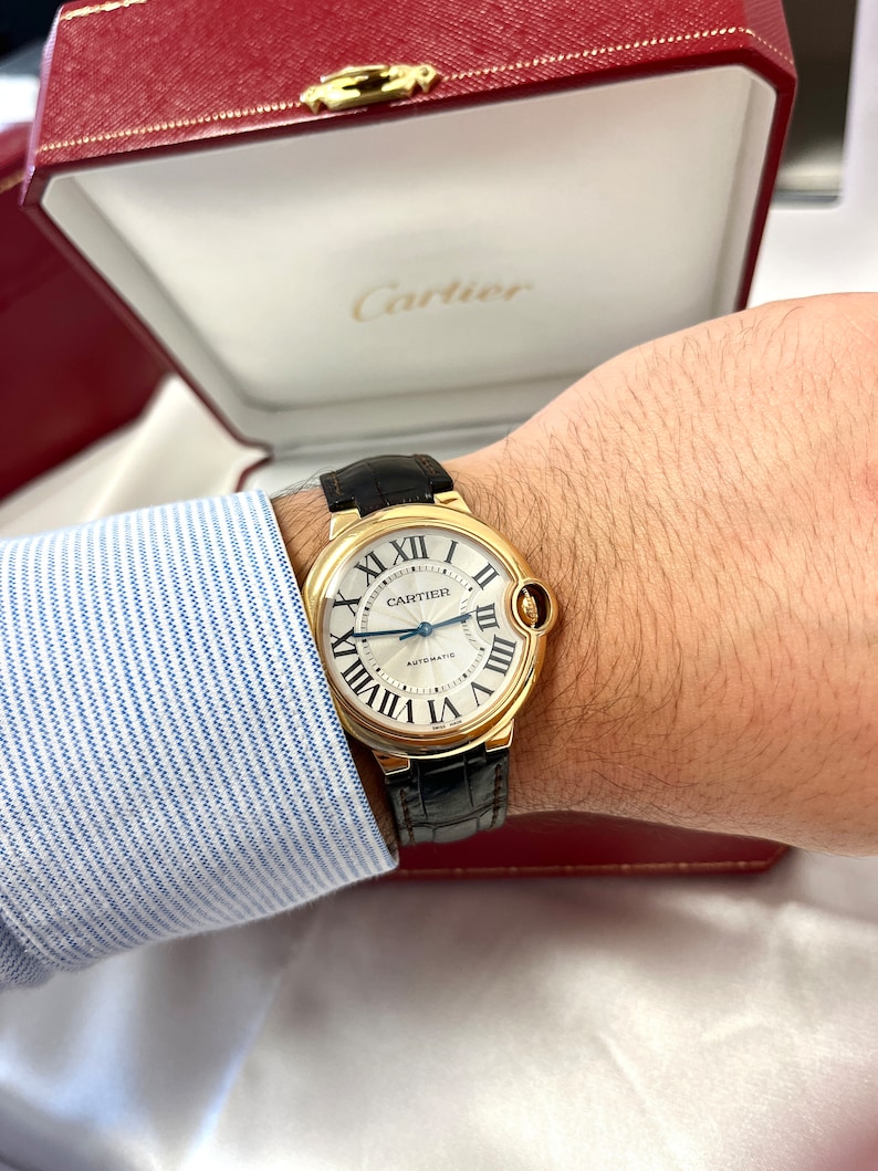 May include: A gold Cartier watch with a white face and black leather band. The watch face has Roman numeral hour markers and a date window at the 3 o'clock position. The watch is labeled 'CARTIER AUTOMATIC'.