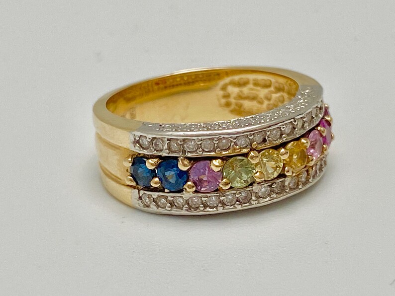 May include: A gold band ring with a row of colorful gemstones set in a white metal band. The gemstones are surrounded by smaller diamonds.