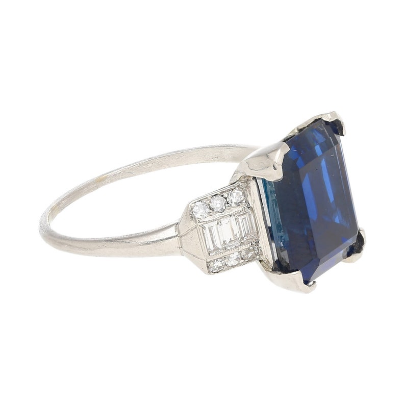 K&ouml;nnte beinhalten: Ein Platinring mit einem gro&szlig;en, tiefblauen Smaragdschliff-Saphir und kleineren Diamanten, die in einem geometrischen Design gefasst sind.