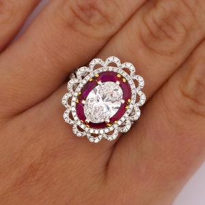 May include: A white gold ring featuring a large oval-shaped diamond surrounded by a halo of smaller diamonds and red rubies. The ring has a delicate, floral design.