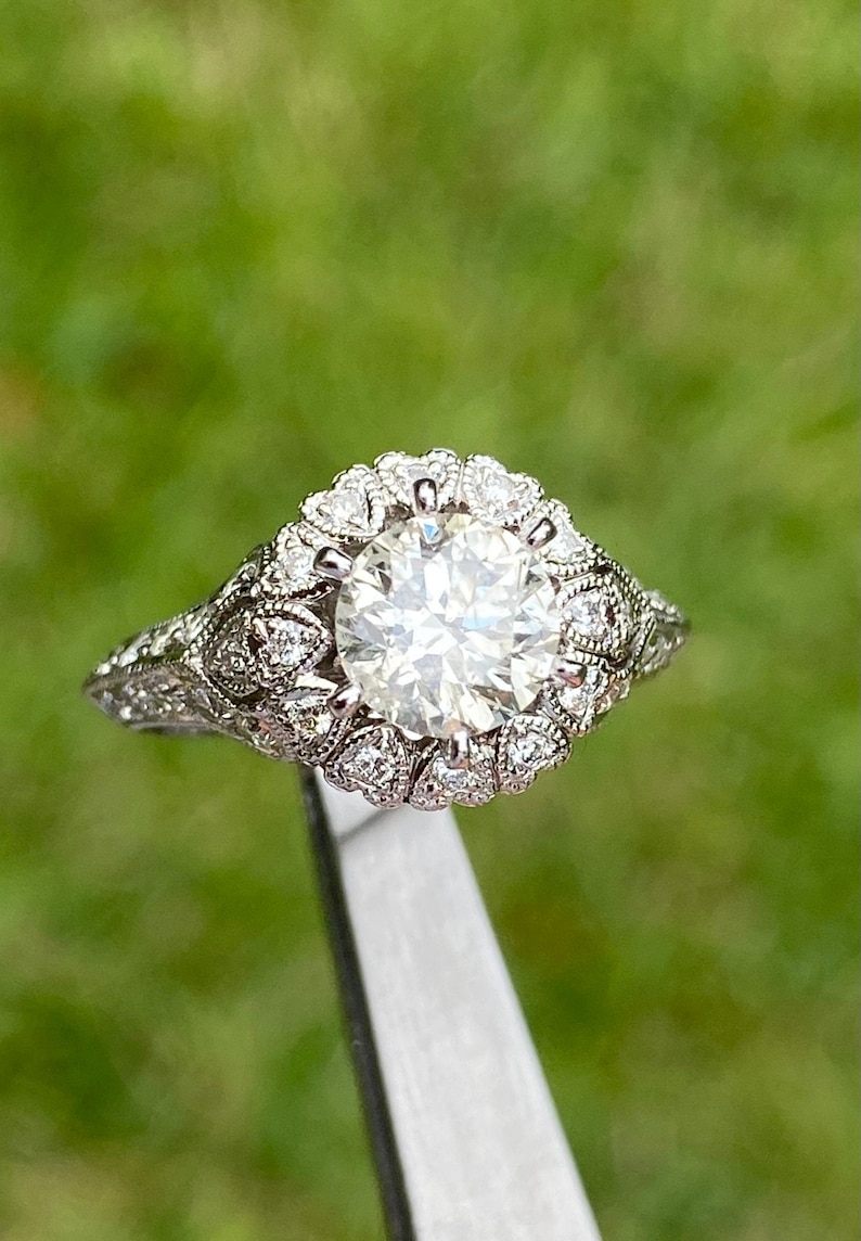 May include: A silver engagement ring with a large, round, clear diamond surrounded by a halo of smaller diamonds. The band is decorated with intricate floral designs.
