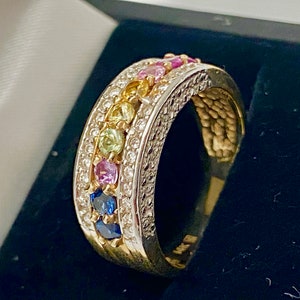 May include: A gold band ring with a row of colorful gemstones set in a white metal band. The gemstones are arranged in a rainbow pattern, with blue, green, yellow, pink, and purple stones.