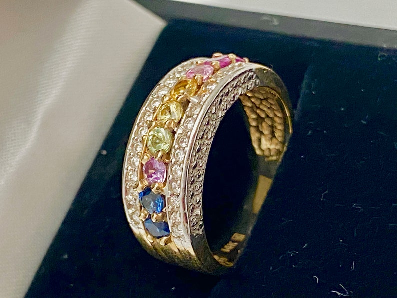 May include: A gold band ring with a row of colorful gemstones set in a white metal band. The gemstones are arranged in a rainbow pattern, with blue, green, yellow, pink, and purple stones.