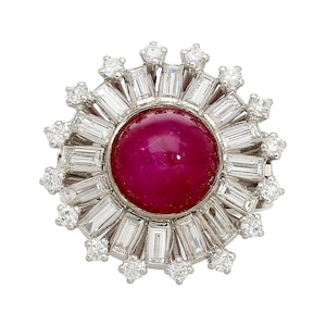 May include: A round, ruby-colored gemstone ring, surrounded by a halo of baguette-cut and round diamonds. The ring's setting is silver-toned, creating a striking contrast with the deep red center stone. The design is reminiscent of a sunburst.