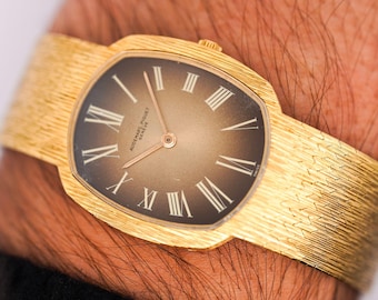 May include: A gold-toned wristwatch with a textured band and a dark brown face. The watch features Roman numeral hour markers and gold hands. The watch is a luxury item.