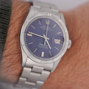 Puede incluir: Un reloj Rolex Oyster Perpetual Date plateado con esfera azul. El reloj presenta el logotipo de Tiffany & Co. y una visualización de la fecha a las 3 en punto. El reloj tiene un brazalete plateado.