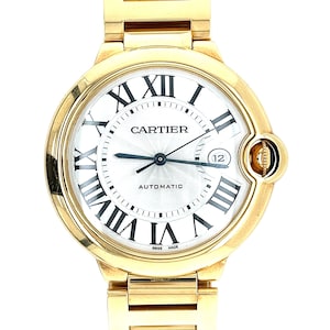 May include: A gold Cartier Ballon Bleu watch with a white face and Roman numeral hour markers. The watch has a date window at the 3 o'clock position and a screw-down crown. The watch is automatic and has a Swiss made inscription.