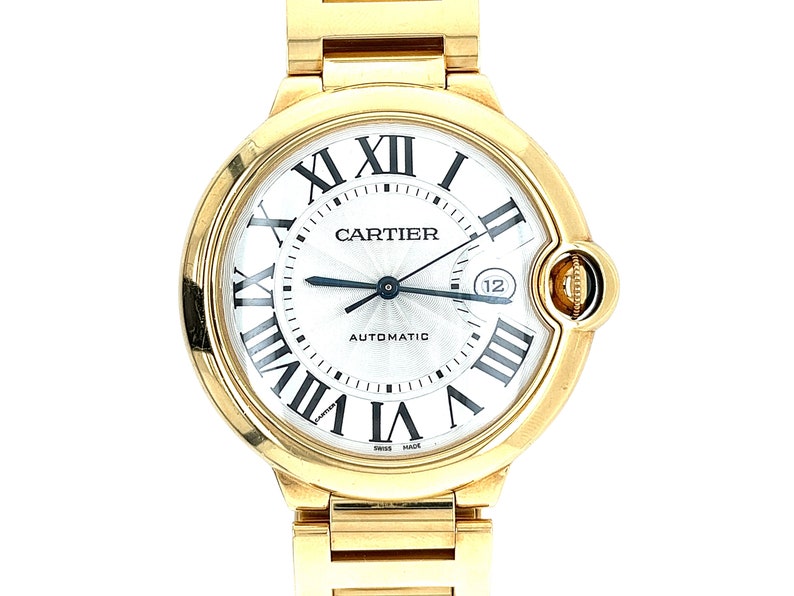 May include: A gold Cartier Ballon Bleu watch with a white face and Roman numeral hour markers. The watch has a date window at the 3 o'clock position and a screw-down crown. The watch is automatic and has a Swiss made inscription.