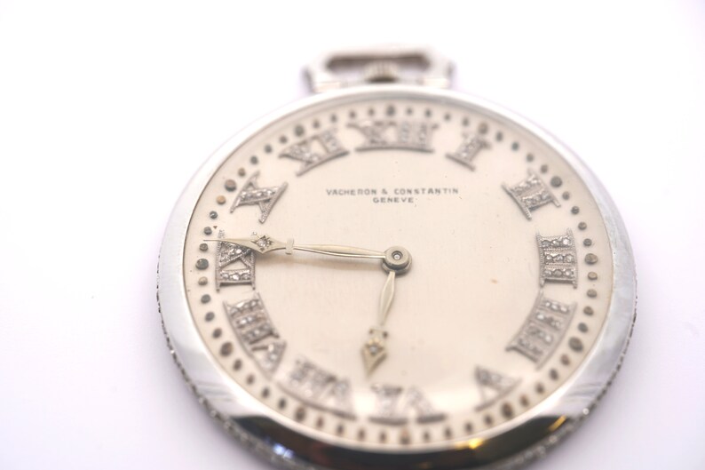 May include: A silver pocket watch with a white face and diamond-encrusted Roman numeral hour markers. The watch face also features the text "VACHERON & CONSTANTIN GENEVE".