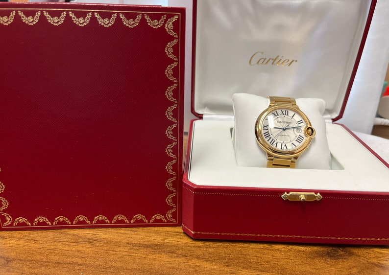 May include: A gold Cartier watch with a white face and Roman numeral hour markers. The watch is in a red box with the Cartier logo on the top.